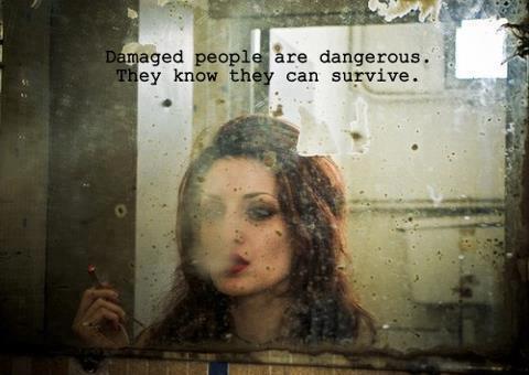 A grainy, weathered photo of a woman with dark tousled hair and heavy eye makeup, looking at herself in a dirty, spotted mirror while holding a lipstick to her lips. Text at the top reads: "Damaged people are dangerous. They know they can survive."