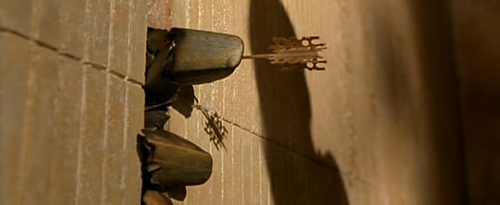 Wide, cinematic film still of a tan, weathered wall with vertical lines. A feathered arrow is lodged in the wall beside rough stone-like chunks, with a thin cord trailing down to a small ornate object. A long dark shadow falls across the wall. No text is visible.