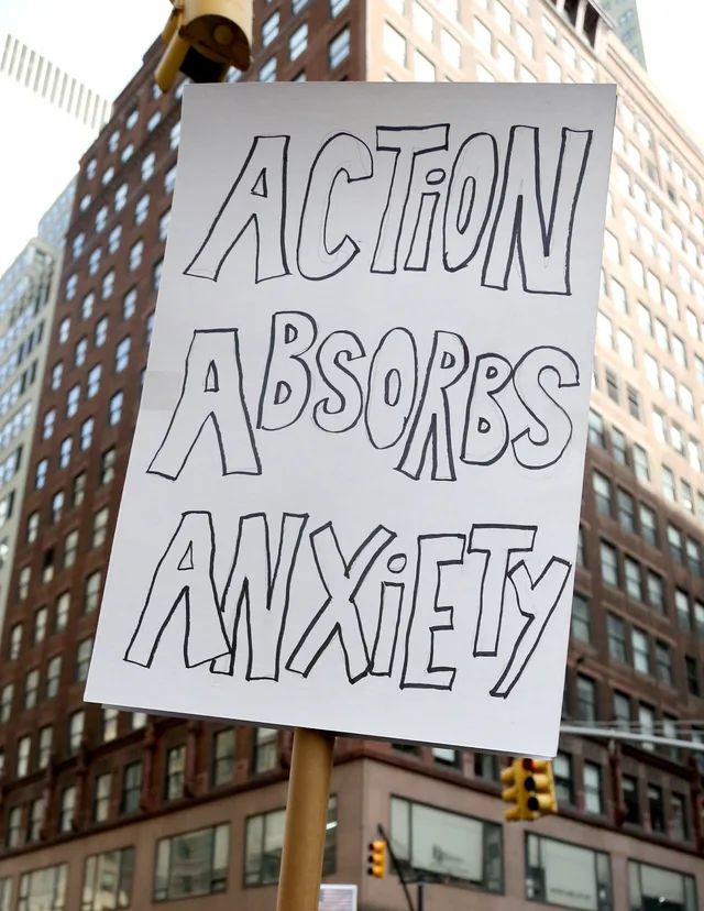 A white poster board on a wooden stick, held up on a city street in front of a tall brick building and traffic lights. Hand-drawn black outline letters read: "ACTION ABSORBS ANXIETY".