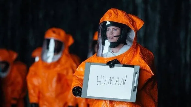 A woman in an orange hazmat suit with a clear helmet and headset holds a clipboard with a white sheet that reads "HUMAN" in handwritten capital letters. Other people in orange hazmat suits stand blurred behind her in a dark setting.
