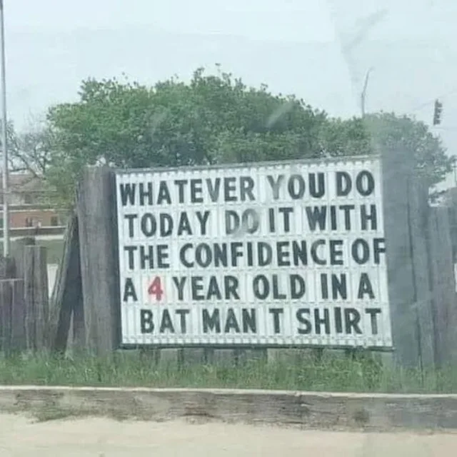 A photo of a white marquee sign with black letters, taken through a smudged window, in front of green trees. It reads: "WHATEVER YOU DO TODAY DO IT WITH THE CONFIDENCE OF A 4 YEAR OLD IN A BAT MAN T SHIRT". The "4" is red.