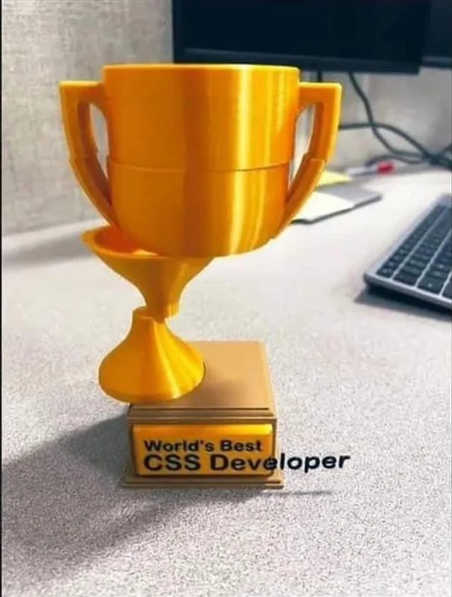 A glossy gold 3D-printed trophy sits on a gray office desk beside a keyboard and monitor. The trophy's pieces are visibly misaligned. The plaque on its base reads "World's Best CSS Developer", and the word "Developer" spills off the plaque onto the desk.