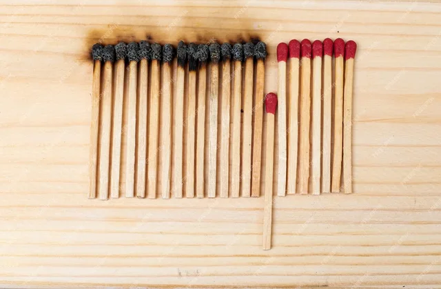 Overhead photo on a light wood surface: fourteen burnt matches with blackened heads in a row on the left, seven unused red-headed matches in a row on the right, and one unused red-headed match set lower, between the two groups. The image has no text.