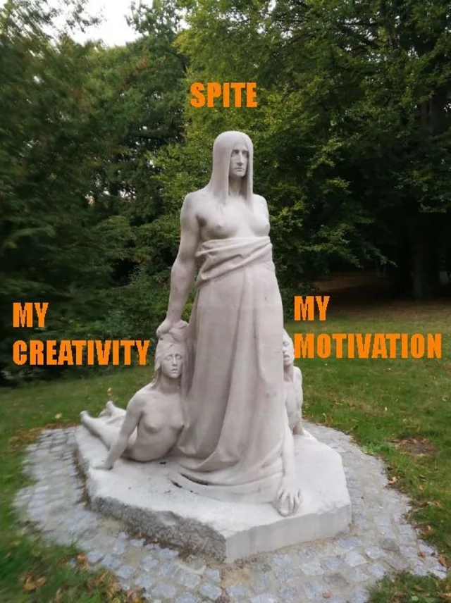 A pale stone statue in a park of a tall robed figure standing over two smaller nude figures, one reclining at left, one crouching at right. Orange text labels the tall figure "SPITE", the left figure "MY CREATIVITY" and the right figure "MY MOTIVATION".