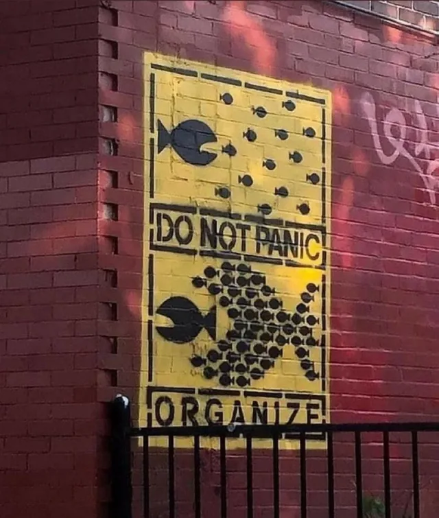 Yellow stencil mural on a red brick wall. At top, a large black fish chases a scattered group of tiny black fish. A middle banner reads "DO NOT PANIC". Below, the small fish are packed into the shape of one giant fish chasing the big fish. A bottom banner reads "ORGANIZE".