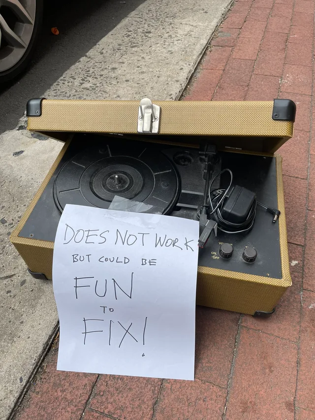 A tweed-covered portable turntable with its lid open sits on a brick sidewalk next to a curb, its power adapter coiled on top. A handwritten paper sign taped to it reads: "DOES NOT WORK BUT COULD BE FUN TO FIX!"