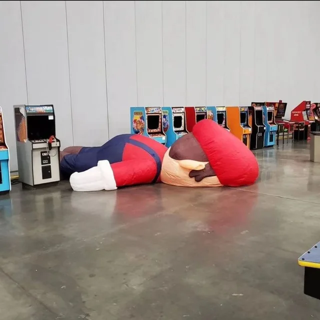 A giant deflated inflatable Mario lies face-down on a concrete warehouse floor, his red cap and white glove crumpled, in front of a row of dark, unpowered vintage arcade cabinets and pinball machines along a gray wall. No text is visible.