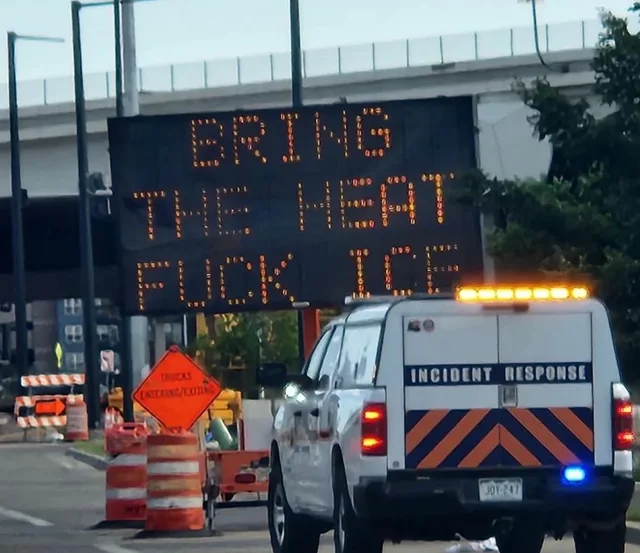 A large roadside electronic message board with orange dot-matrix lettering reads "BRING THE HEAT FUCK ICE". Below it sit orange construction barrels, a diamond sign reading "Trucks Entering/Exiting", and a white truck with flashing lights labeled "INCIDENT RESPONSE".