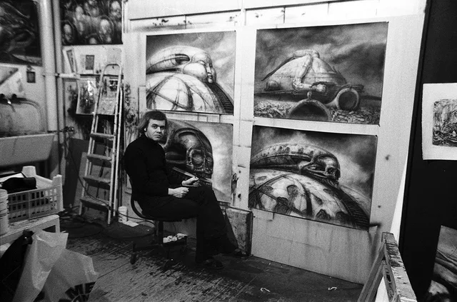 Black-and-white photo of a man with shaggy hair in a black turtleneck, seated on a stool in a cluttered studio. Behind him, four large airbrushed paintings show biomechanical domed structures with skulls and faces on rails. No text.