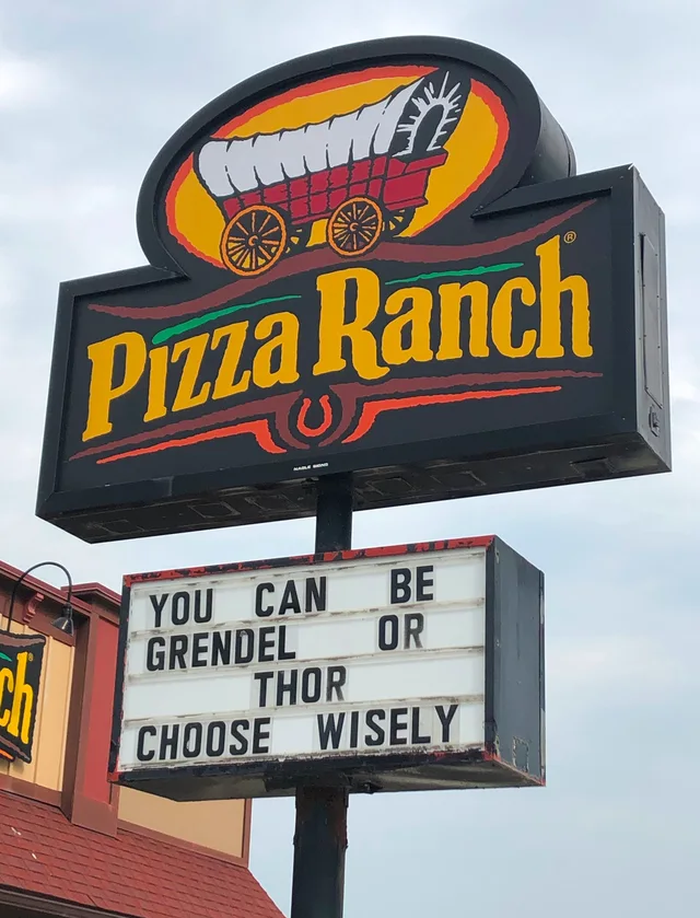 A tall roadside Pizza Ranch sign with a covered wagon logo and yellow lettering on a black background. Below it, a marquee with black letters reads: "YOU CAN BE GRENDEL OR THOR. CHOOSE WISELY". A cloudy sky is behind it.
