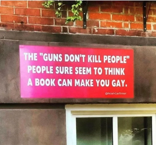 A pink-to-red sign mounted on a dark stone wall below a red brick wall, above a white-framed window. White text reads: "THE "GUNS DON'T KILL PEOPLE" PEOPLE SURE SEEM TO THINK A BOOK CAN MAKE YOU GAY." Small text at bottom right: "@NoahGarfinkel".