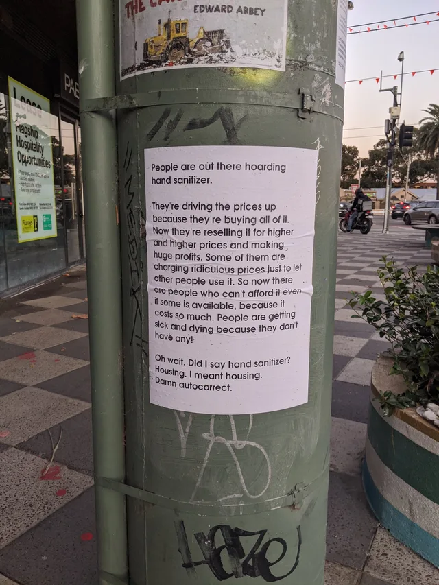 A white paper flyer pasted on a green metal street pole, with graffiti around it. Text: "People are out there hoarding hand sanitizer. They're driving the prices up because they're buying all of it. Now they're reselling it for higher and higher prices and making huge profits. Some of them are charging ridiculous prices just to let other people use it. So now there are people who can't afford it even if some is…
