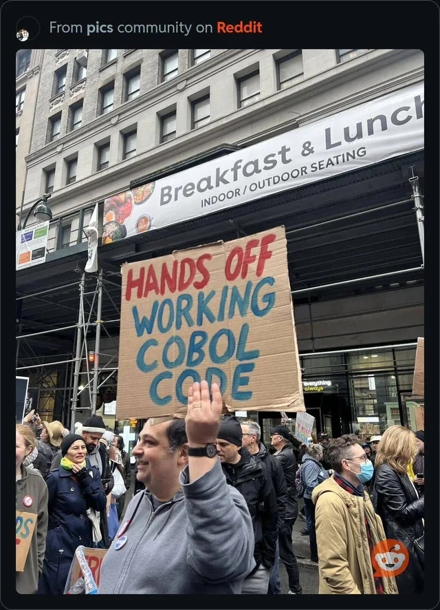 Reddit post from the pics community. A man in a gray hoodie raises one hand in a street crowd, holding a cardboard sign hand-lettered in red and blue: "HANDS OFF WORKING COBOL CODE". Behind him a banner reads "Breakfast & Lunch INDOOR / OUTDOOR SEATING".