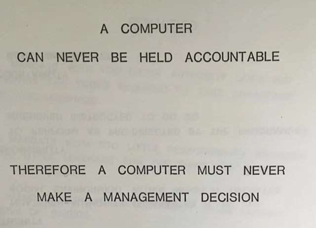 Photo of a printed page in black capital letters that reads: "A COMPUTER CAN NEVER BE HELD ACCOUNTABLE" followed by "THEREFORE A COMPUTER MUST NEVER MAKE A MANAGEMENT DECISION". Faint mirrored text from the reverse side shows through the paper.