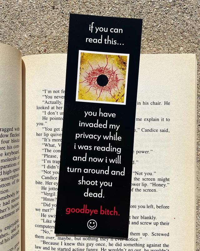 A black bookmark lying on an open paperback. White text: "if you can read this..." Below is a square photo of a bloodshot yellow eye with a black pupil. More text: "you have invaded my privacy while i was reading and now i will turn around and shoot you dead." Red text: "goodbye bitch." A white smiley face is at the bottom.