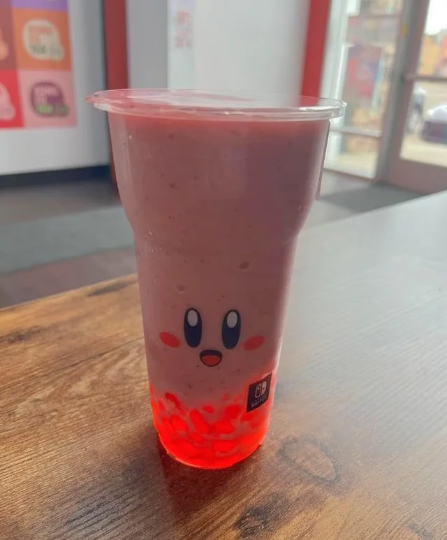 A clear plastic cup holds a pink fruit smoothie on a wooden table, with red boba pearls in the bottom. A cute face is printed on the cup: two tall blue oval eyes with white highlights, round pink cheeks, and a small red mouth. A small Nintendo Switch logo sits near the base.