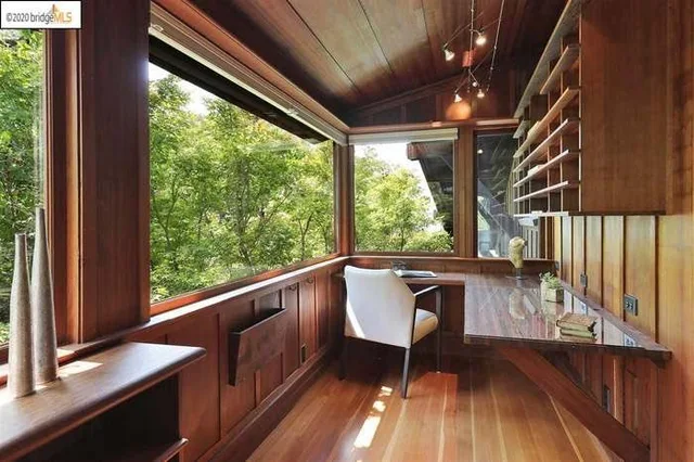 A small wood-paneled writing room with a built-in desk along the wall, a white chair, and large windows looking out at green trees. Built-in shelves hang on the right, and two tall vases sit on the left. Corner text reads: 2020 bridgeMLS.
