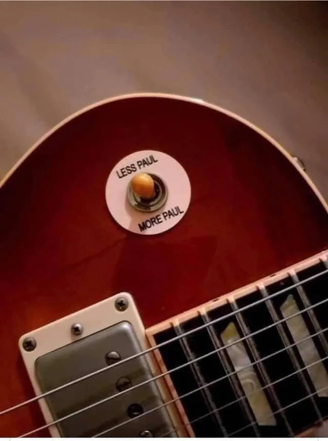 Close-up of a sunburst red Gibson Les Paul style electric guitar. On the upper body, a round white sticker around the toggle switch reads "LESS PAUL" at the top and "MORE PAUL" at the bottom. The switch has an amber tip.