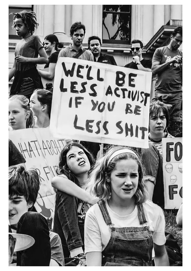 Black-and-white photo of young protesters sitting and standing outside a columned building. A girl in a headband and denim overalls squints in front, while a girl behind her grimaces. A large handmade sign reads: "WE'LL BE LESS ACTIVIST IF YOU BE LESS SHIT". Partial signs nearby read "WHAT ABOUT", "FUT" and "FO".