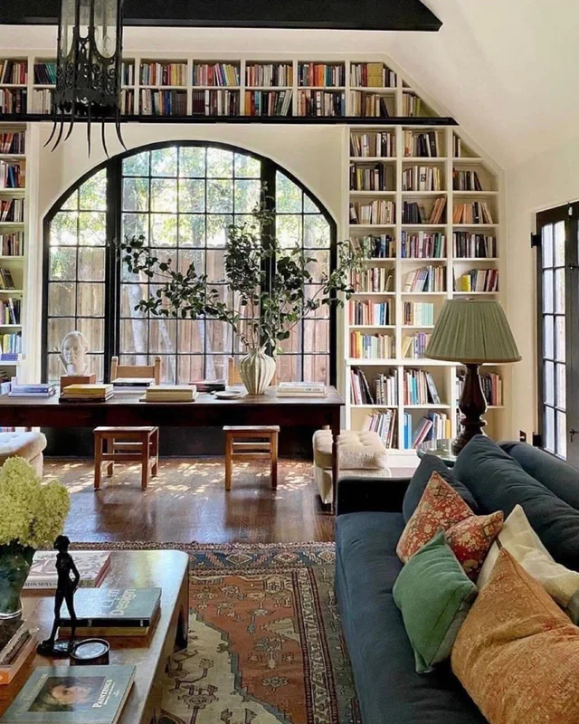 A sunlit living room with floor-to-ceiling white built-in bookshelves, a high gallery of books under a vaulted ceiling, and a large black-framed arched window. A long wooden table with stacked books and a plant stands in front of it, with a navy sofa, colorful pillows, a patterned rug and a lamp. No text.