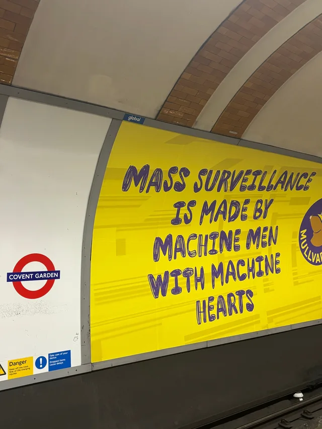 Photo of a curved yellow poster on a London Underground platform at Covent Garden, beside a red-and-blue roundel sign reading "COVENT GARDEN". Purple scribbly hand-lettered text on the poster reads: "MASS SURVEILLANCE IS MADE BY MACHINE MEN WITH MACHINE HEARTS".