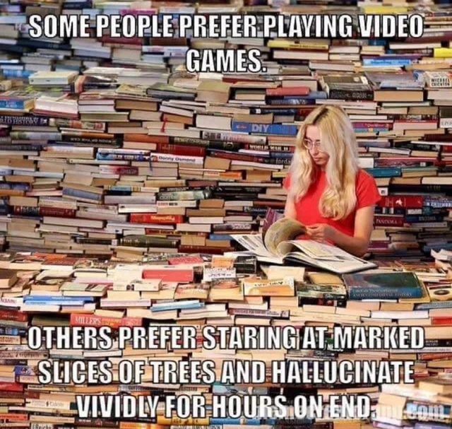 A blonde woman with glasses in a red t-shirt sits surrounded by enormous towering piles of books, reading a large open book. Top text: "SOME PEOPLE PREFER PLAYING VIDEO GAMES." Bottom text: "OTHERS PREFER STARING AT MARKED SLICES OF TREES AND HALLUCINATE VIVIDLY FOR HOURS ON END."