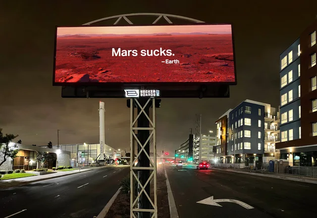 A lit LED billboard at night shows a red Martian desert landscape with white text reading "Mars sucks." and below it "—Earth". The sign reads "Becker Boards.com". Below, an empty street, apartment buildings, and a tall white rocket labeled SpaceX stand in the background.