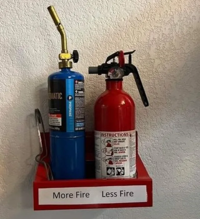 A red wall-mounted shelf holding a blue propane torch cylinder with a brass nozzle on the left and a red fire extinguisher with an instructions label on the right. A white label along the front edge reads: "More Fire Less Fire".