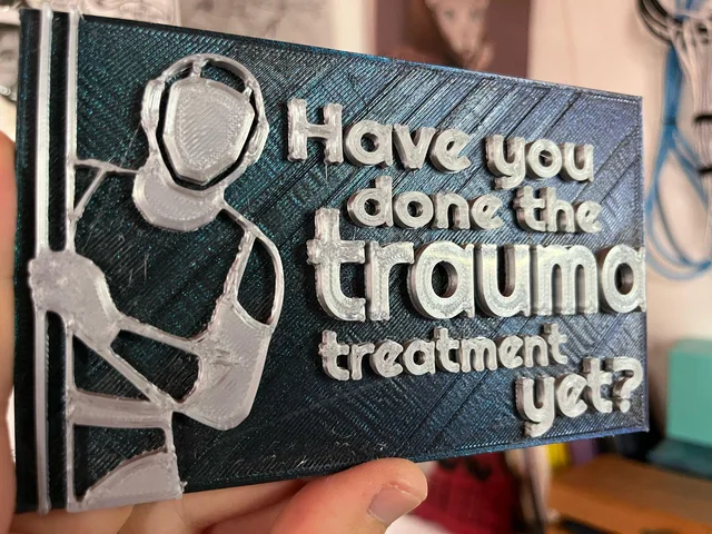 A 3D-printed plaque with raised silver filament on a shimmery dark blue-green base, held in a hand. On the left is an outlined figure in a helmet and headset, gripping a vertical pole. Text on the right reads: "Have you done the trauma treatment yet?"