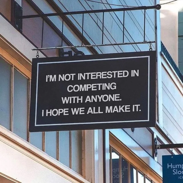 A black hanging shop sign with a white border, suspended from a metal bracket outside a building. White capital letters read: "I'M NOT INTERESTED IN COMPETING WITH ANYONE. I HOPE WE ALL MAKE IT." A blue sign partly reading "Hump" and "Sloo" is visible at the lower right.