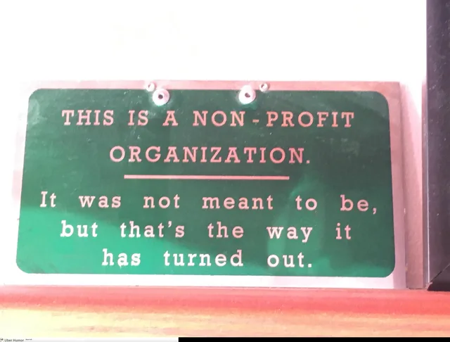 Photo of a green metal sign with engraved lettering, mounted on a shelf. It reads: "THIS IS A NON-PROFIT ORGANIZATION." A line follows, then: "It was not meant to be, but that's the way it has turned out." A small Uber Humor watermark is in the corner.