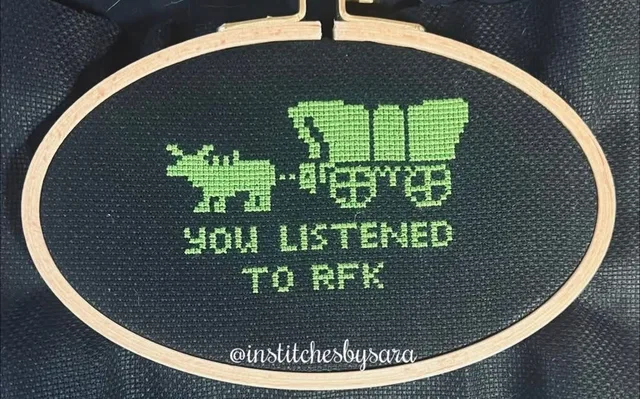 Green cross-stitch on black fabric in an oval wooden embroidery hoop. It shows the pixelated ox and covered wagon from The Oregon Trail. Beneath it, stitched text reads: "YOU LISTENED TO RFK". A watermark reads: "@institchesbysara".