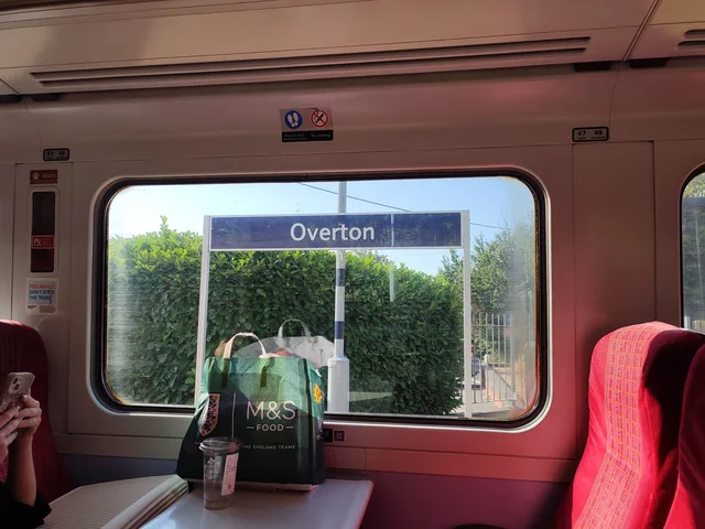 Photo from inside a train carriage looking through a window at a platform sign reading "Overton" in white letters on a dark blue board, in front of a green hedge. On the table sits a green M&S Food bag reading "M&S FOOD" and "THE ENGLAND TEAMS", plus a plastic cup. Red seats flank the window.