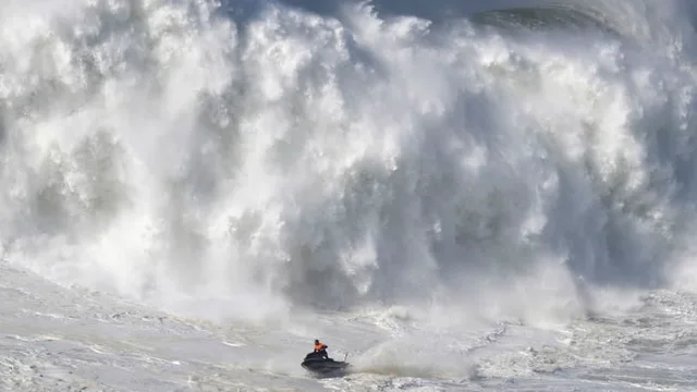 Photo of a huge wall of white churning surf and foam towering over a tiny person in an orange vest riding a dark jet ski on the choppy water below. There is no text in the image.