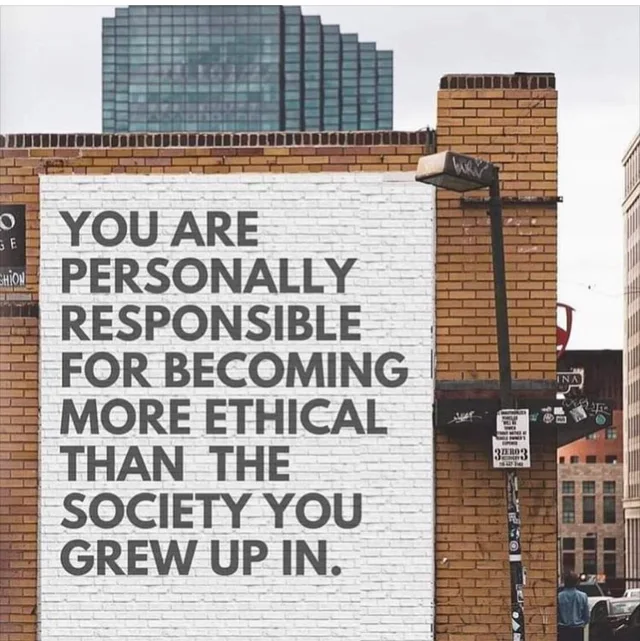 A white-painted section of a brick wall on a city street displays a mural in large gray capital letters: "YOU ARE PERSONALLY RESPONSIBLE FOR BECOMING MORE ETHICAL THAN THE SOCIETY YOU GREW UP IN." A street lamp, tall glass building, and posted signs are visible around it.