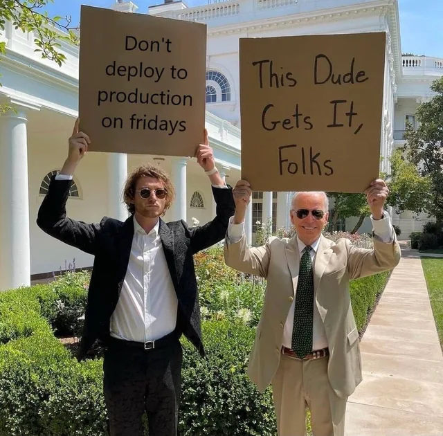 Two men in sunglasses stand outdoors in front of a white building, each holding a cardboard sign overhead. The younger man in a black jacket holds a sign reading "Don't deploy to production on fridays". The older man in a tan suit and green tie holds a sign reading "This Dude Gets It, Folks".
