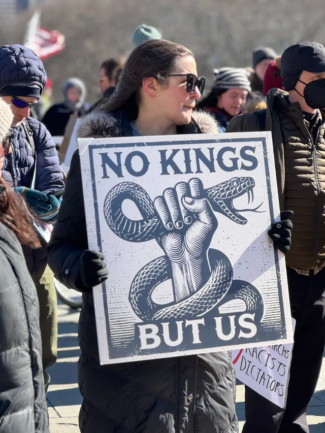 A woman in sunglasses and a fur-trimmed black coat holds a white protest sign in a crowd. The sign has a blue woodcut-style illustration of a raised fist strangling a coiled snake with open fangs. Text reads: "NO KINGS" at the top and "BUT US" at the bottom.