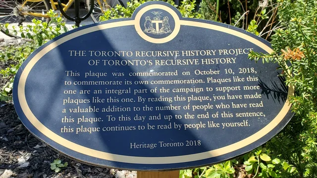 Oval blue and gold heritage plaque with a city crest, in a garden. Text: "THE TORONTO RECURSIVE HISTORY PROJECT OF TORONTO'S RECURSIVE HISTORY. This plaque was commemorated on October 10, 2018, to commemorate its own commemoration. Plaques like this one are an integral part of the campaign to support more plaques like this one. By reading this plaque, you have made a valuable addition to the number of people who…