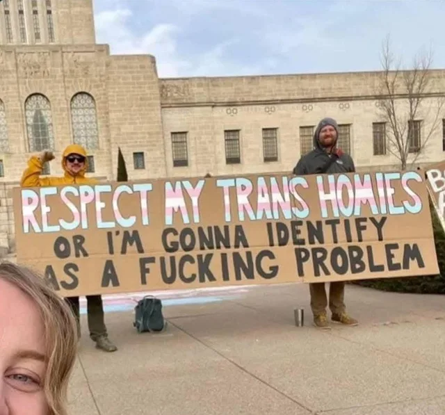 Two bearded people in winter jackets, one in a yellow hooded raincoat with a raised fist, hold a huge cardboard banner outside a stone government building. Trans flag colored letters read: "RESPECT MY TRANS HOMIES" then in black: "OR I'M GONNA IDENTIFY AS A FUCKING PROBLEM". Part of a face is in the corner.