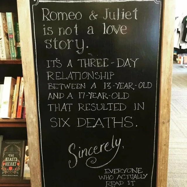 A chalkboard sandwich sign in front of bookshelves. Text: "Romeo & Juliet is not a love story. IT'S A THREE-DAY RELATIONSHIP BETWEEN A 13-YEAR-OLD AND A 17-YEAR-OLD THAT RESULTED IN SIX DEATHS. Sincerely, EVERYONE WHO ACTUALLY READ IT"