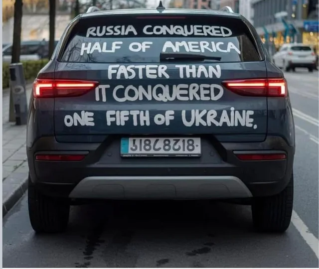 Rear view of a dark gray SUV parked on a city street, with hand-painted white capital letters across the back window and tailgate that read: "RUSSIA CONQUERED HALF OF AMERICA FASTER THAN IT CONQUERED ONE FIFTH OF UKRAINE." The license plate is mirror-reversed.
