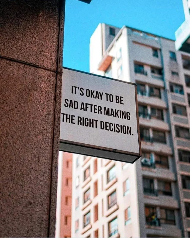A white square sign mounted on a pinkish granite wall, with a blue sky and tall apartment buildings behind it. The sign reads: "IT'S OKAY TO BE SAD AFTER MAKING THE RIGHT DECISION."