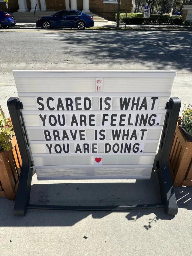 A white sidewalk letterboard sign on a black stand, between two wooden planters, with black letters reading: "SCARED IS WHAT YOU ARE FEELING. BRAVE IS WHAT YOU ARE DOING." A red heart sits below the text. A street and a parked blue car are behind it.