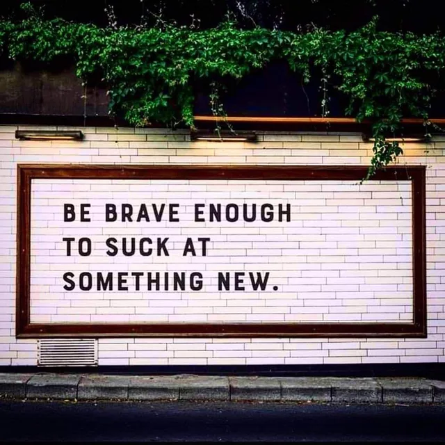 A white subway-tile wall with a wooden-framed panel lit by lamps and draped with green ivy above. Black capital letters read: "BE BRAVE ENOUGH TO SUCK AT SOMETHING NEW." A sidewalk curb and dark street run below.