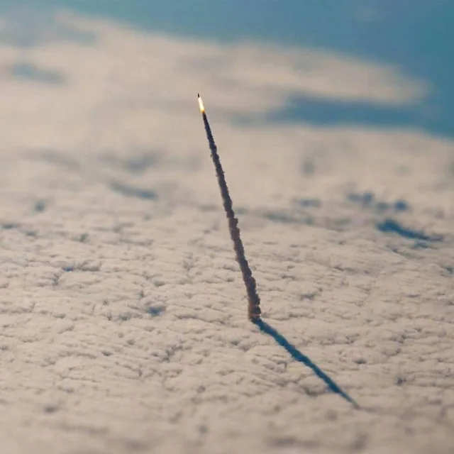 Aerial view of a thick layer of textured white clouds with a thin dark smoke trail rising at a slant, a bright glowing flame at its tip, and a long blue shadow of the trail stretching across the clouds. Blue sky is at the top. No text.