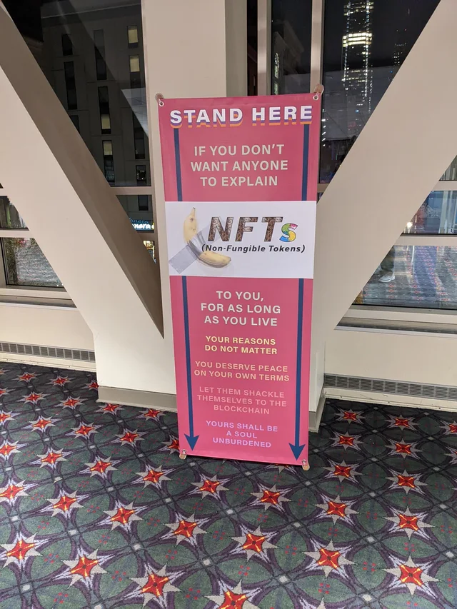 Photo of a pink standing banner in a convention center hall, in front of a large white V-shaped column. Text: "STAND HERE" "IF YOU DON'T WANT ANYONE TO EXPLAIN" then a white band with a duct-taped banana beside "NFTs (Non-Fungible Tokens)" then "TO YOU, FOR AS LONG AS YOU LIVE" "YOUR REASONS DO NOT MATTER" "YOU DESERVE PEACE ON YOUR OWN TERMS" "LET THEM SHACKLE THEMSELVES TO THE BLOCKCHAIN" "YOURS SHALL BE A SOUL…