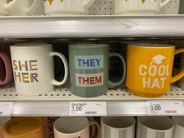 Store shelf photo of three $3.00 mugs. A white mug reads "SHE HER" in colorful striped letters. A green mug reads "THEY" and "THEM" in two labeled boxes. A yellow mug shows a graduation cap and reads "COOL HAT". Shelf tags read "Room Essntls They Them Mug 3.00" and "RE One Day Mug 3.00".