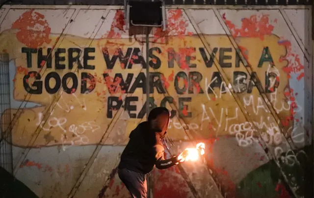 At night, a hooded figure in dark clothing crouches in front of a weathered street mural, holding a flaming object that casts an orange glow. The mural, splattered with red paint and white graffiti, reads in black capital letters: "THERE WAS NEVER A GOOD WAR OR A BAD PEACE".