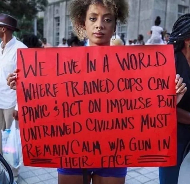 A young woman with curly hair, hoop earrings and a nose ring stands in an outdoor crowd holding a red poster board with handwritten black marker text: "We live in a world where trained cops can panic & act on impulse but untrained civilians must remain calm w/a gun in their face". The last line is underlined.
