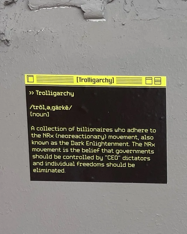 A sticker on a gray, peeling wall styled like a retro computer window with a yellow title bar reading "[Trolligarchy]". Yellow text on black reads: ">> Trolligarchy /trōl,ə,gärkē/ [noun] A collection of billionaires who adhere to the NRx (neoreactionary) movement, also known as the Dark Enlightenment. The NRx movement is the belief that governments should be controlled by "CEO" dictators and individual freedoms…